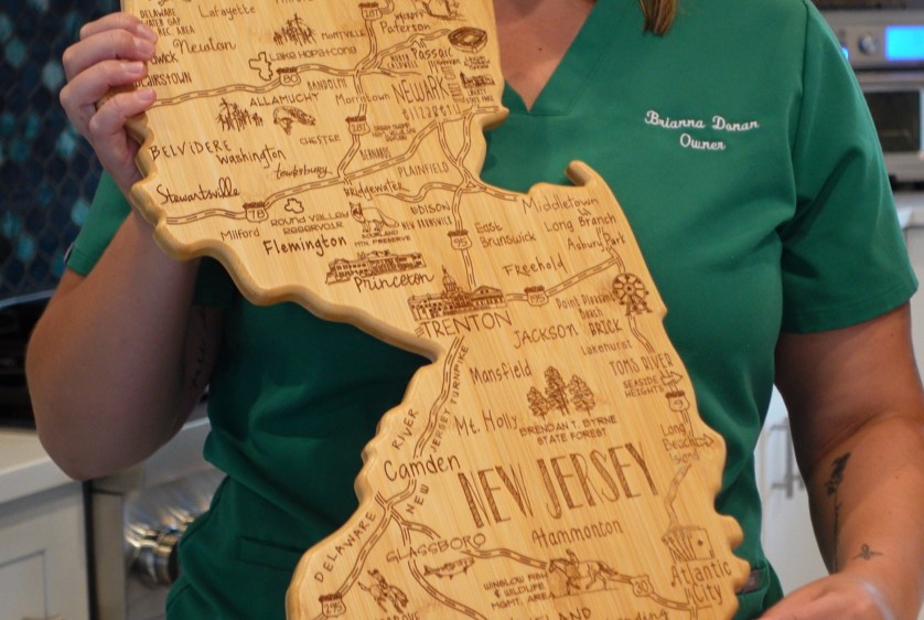 Brianna Donan, owner of Pampered Spirit, holding a wood-engraved New Jersey map, representing travel-inspired massage services across NJ.